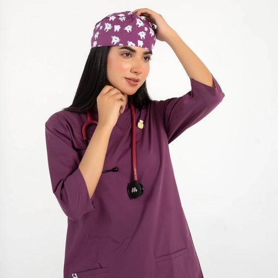 A healthcare professional wearing a purple V-neck medical scrub top, an enamel lapel pin, a stethoscope, and a matching tooth-pattern scrub cap while adjusting her cap against a white background.