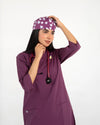 A healthcare professional wearing a purple V-neck medical scrub top, an enamel lapel pin, a stethoscope, and a matching tooth-pattern scrub cap while adjusting her cap against a white background.