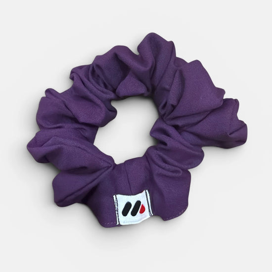 A purple fabric hair scrunchie with a small branded white tag featuring black and red abstract marks against a white background.