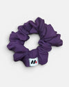A purple fabric hair scrunchie with a small branded white tag featuring black and red abstract marks against a white background.