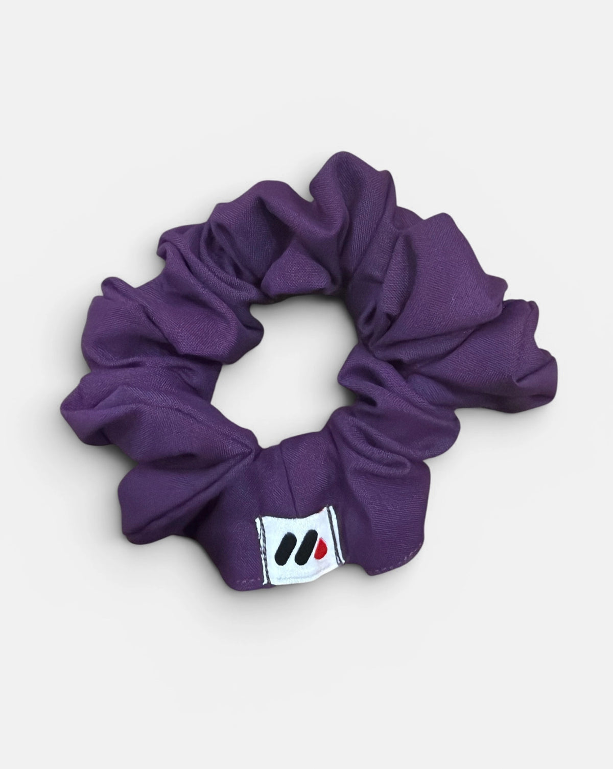 A purple fabric hair scrunchie with a small branded white tag featuring black and red abstract marks against a white background.