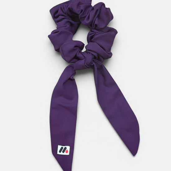A purple fabric hair scrunchie with long tails and a small branded white tag featuring a black and red logo against a white background.