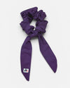 A purple fabric hair scrunchie with long tails and a small branded white tag featuring a black and red logo against a white background.