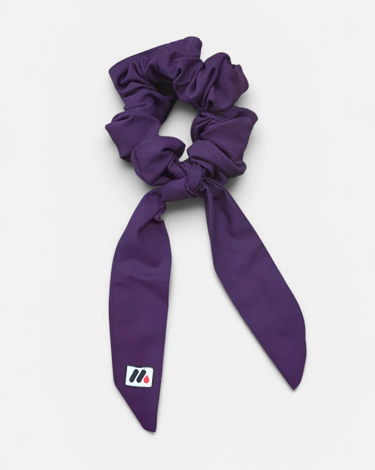 A purple fabric hair scrunchie with long tails and a small branded white tag featuring a black and red logo against a white background.