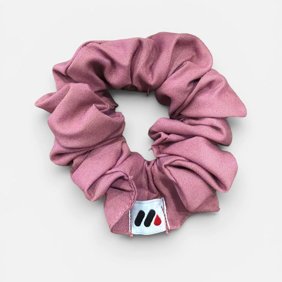 A rose pink fabric hair scrunchie with a small branded white tag featuring black and red abstract marks against a white background.