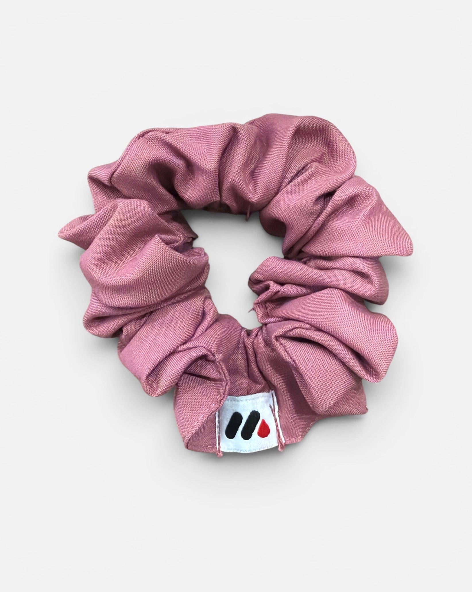 A rose pink fabric hair scrunchie with a small branded white tag featuring black and red abstract marks against a white background.