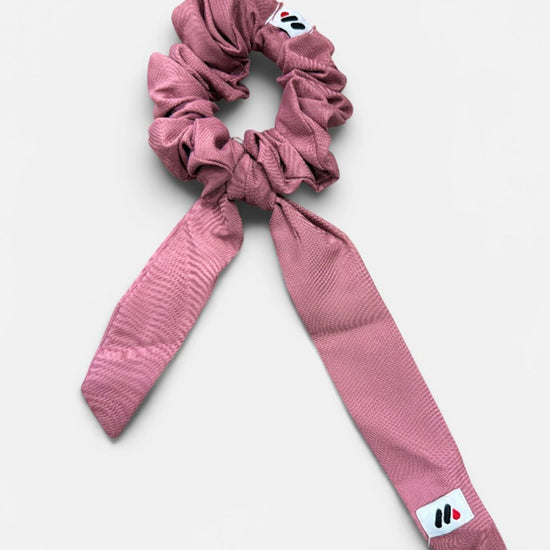 A rose pink fabric hair scrunchie with long tails and a small branded white tag featuring a black and red logo against a white background.