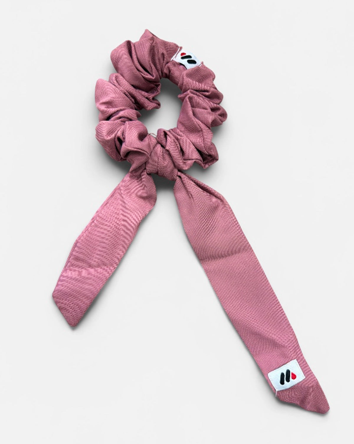 A rose pink fabric hair scrunchie with long tails and a small branded white tag featuring a black and red logo against a white background.