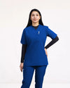 A healthcare professional modeling a matching royal blue V-neck scrub top and pants over a black long-sleeve undershirt against a white studio background.